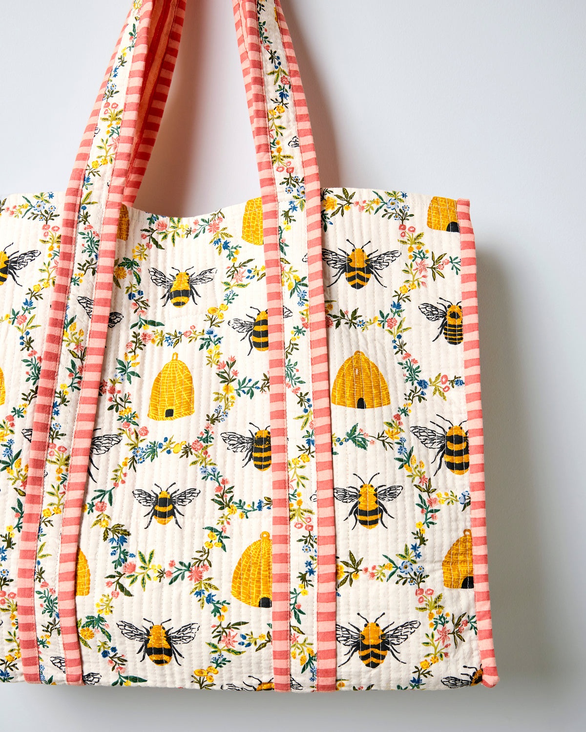 Close view of the cream quilted traveler tote showing bee, beehive, and floral print with pink striped handles and edge trim.