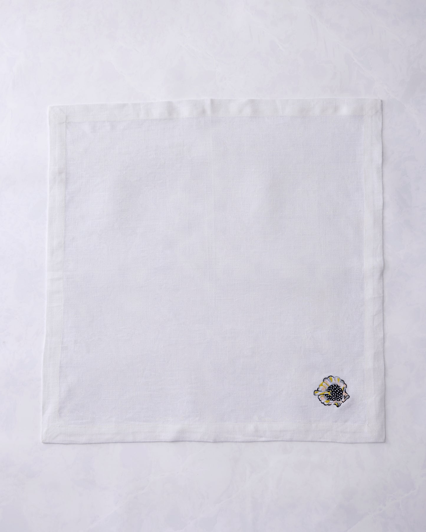 Single white dinner napkin shown flat with an embroidered shell motif near the lower-right corner.
