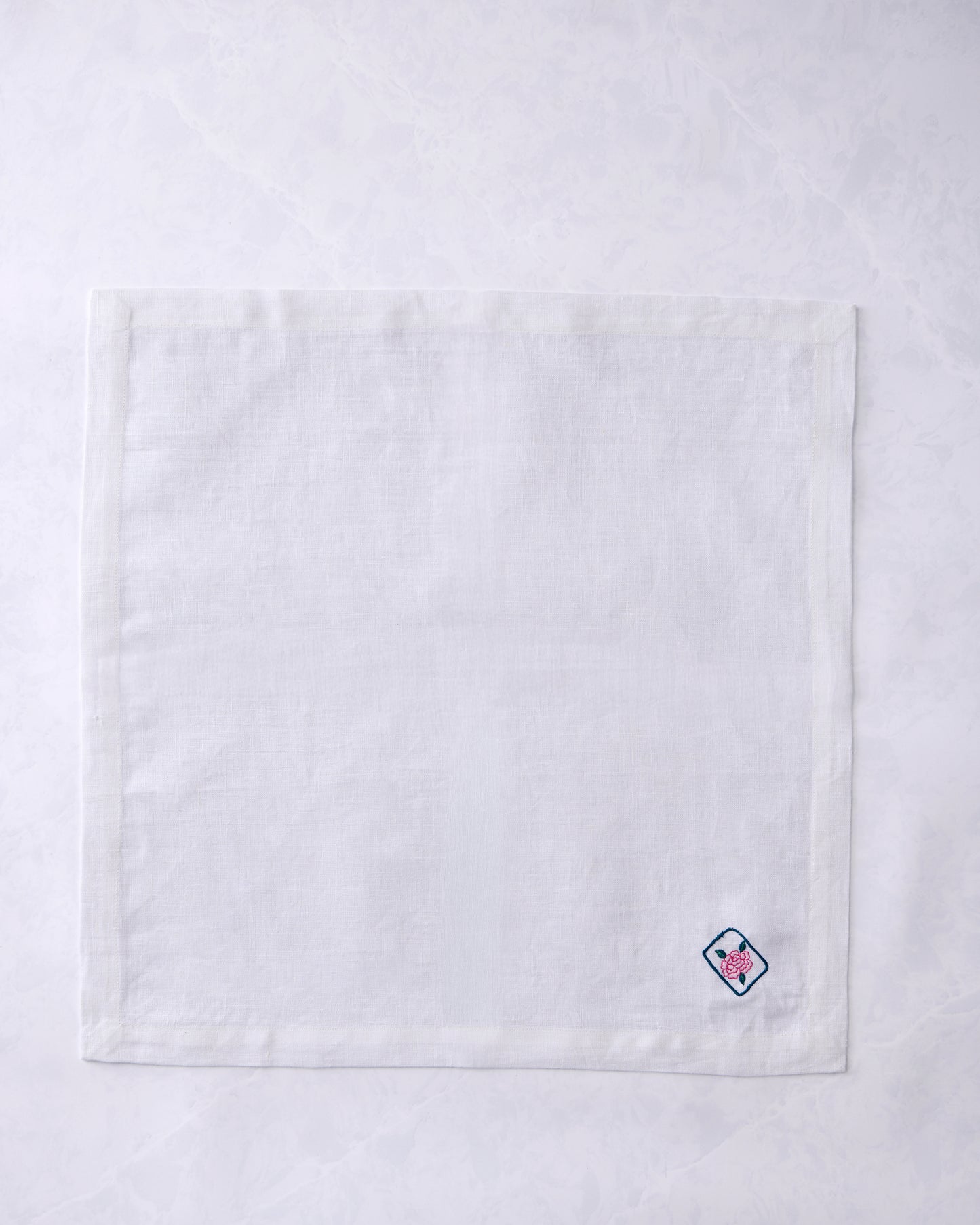 White dinner napkin lying flat with a small embroidered outlined tile containing a pink flower near the lower right corner.