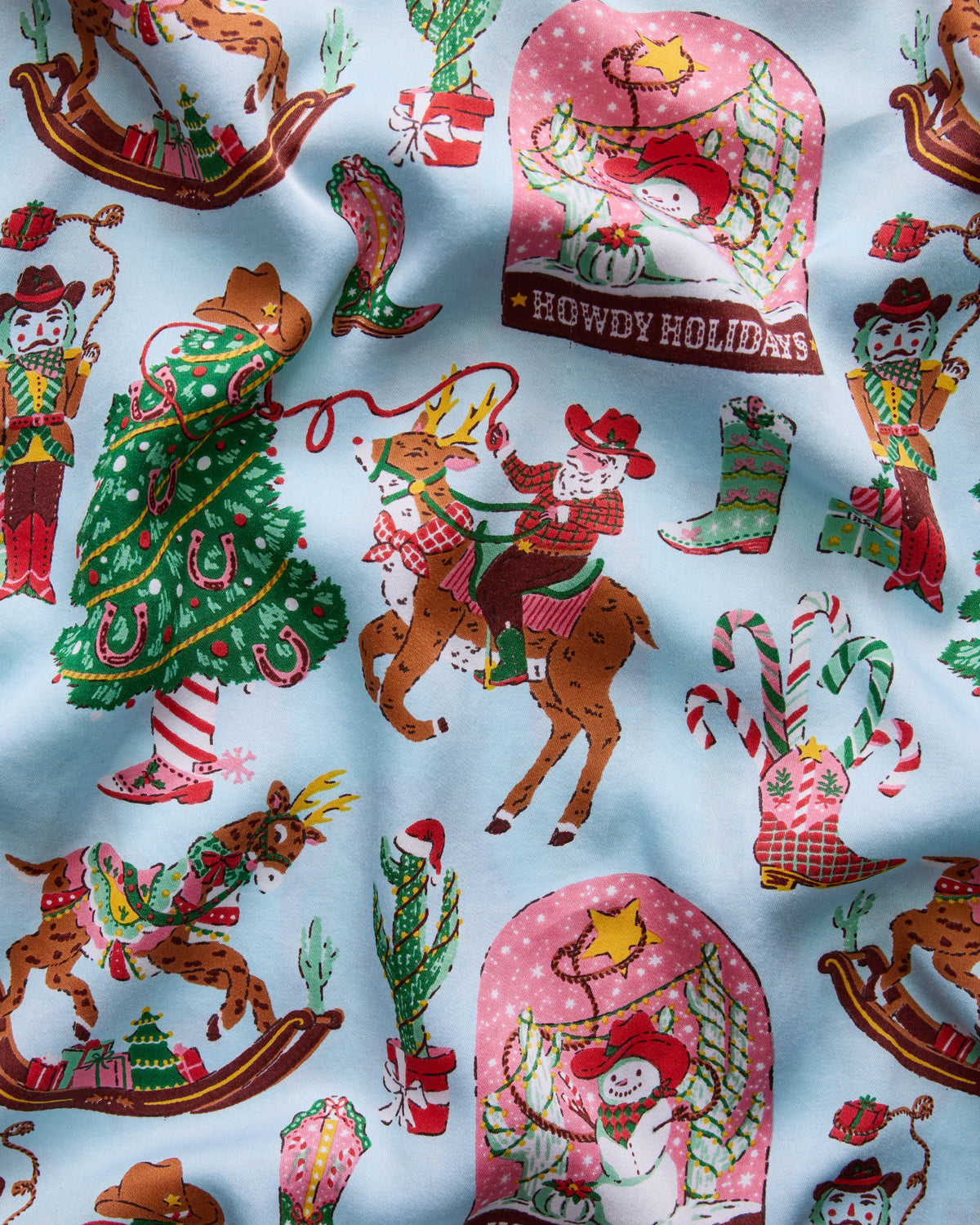Seamless light-blue Cowboy Christmas pajama print with Santa, reindeer, cowboy boots, Christmas trees, candy canes, and a panel reading “HOWDY HOLIDAYS.”