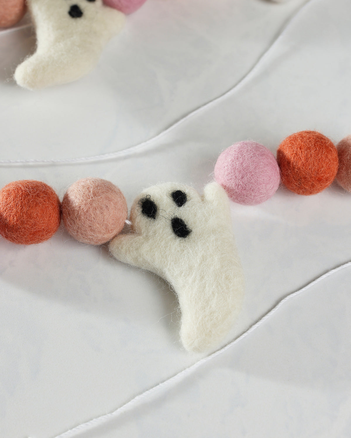 Close-up of a white felt ghost with black facial features between pink and coral felt balls on a garland string.