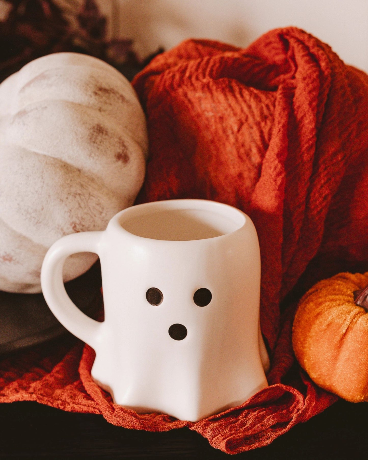 White ghost-shaped mug filled with a light-colored drink, resting on a red blanket beside pumpkins.