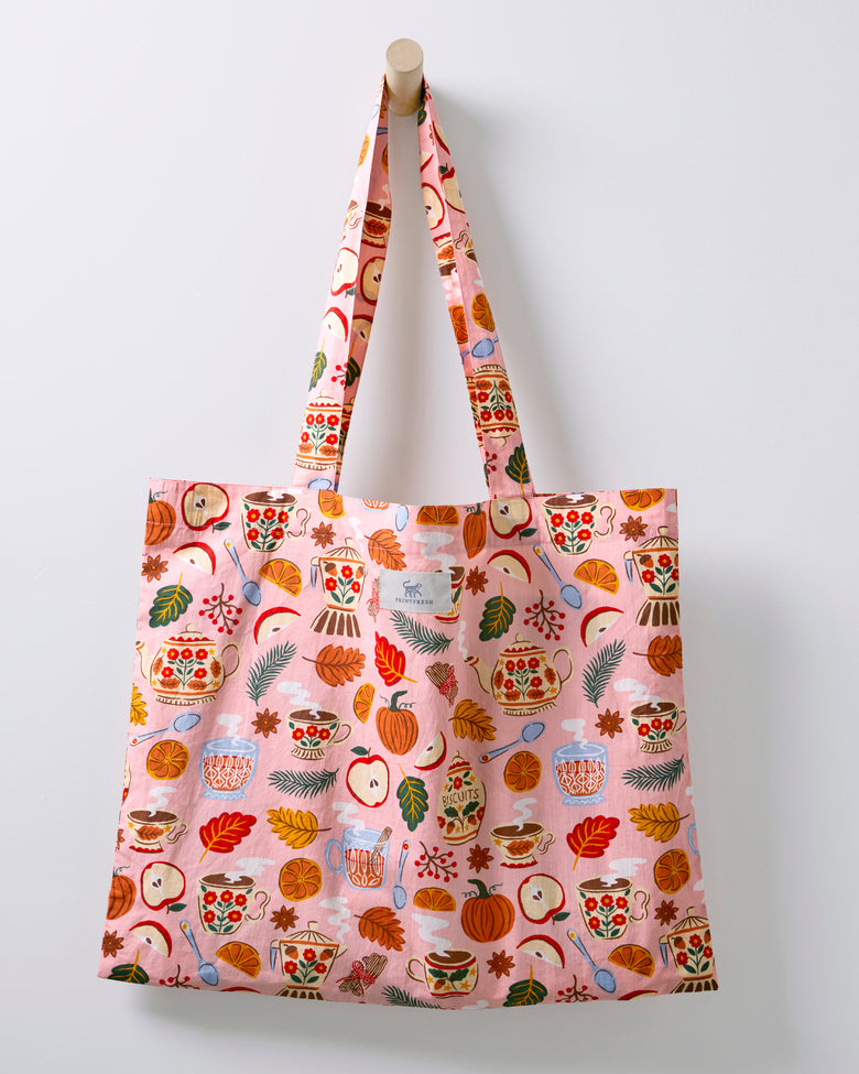 Rose pink tea-themed tote bag hanging from a wooden peg, with long fabric handles.