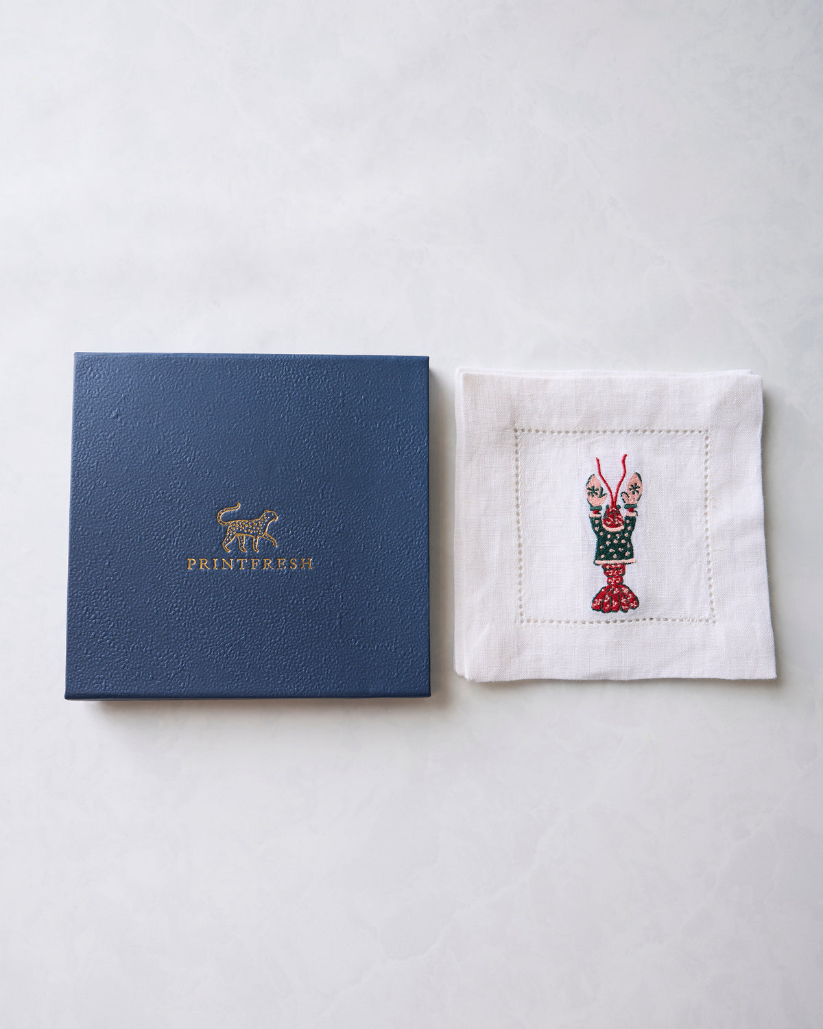 White embroidered cocktail napkin beside a dark blue Printfresh presentation box.