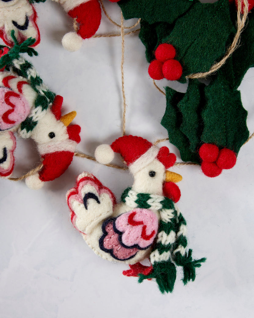 Close-up of a felt hen in a Santa hat beside holly leaves and red berries on the garland.