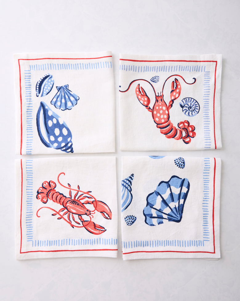 Four folded white Lobster Landing napkins arranged in a square, decorated with blue shells and red lobsters.