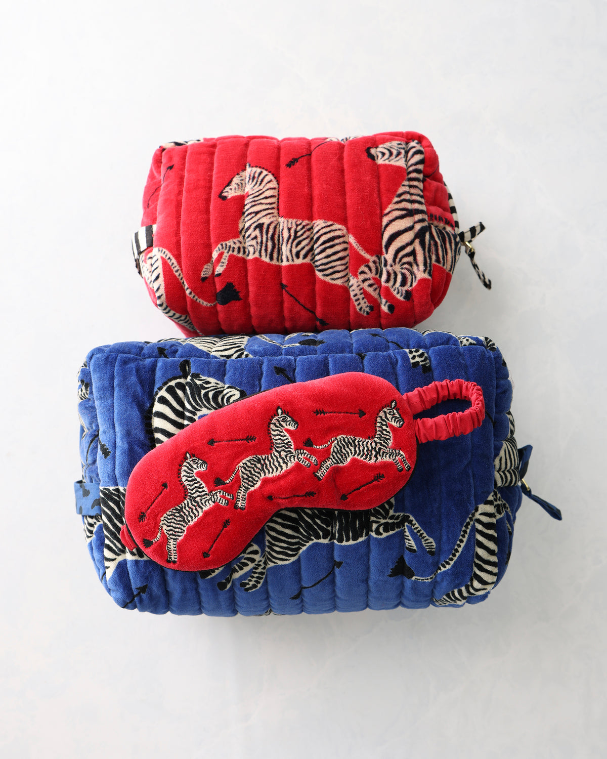 Red zebra-print eye mask resting across a blue quilted pouch, with a red pouch behind it.