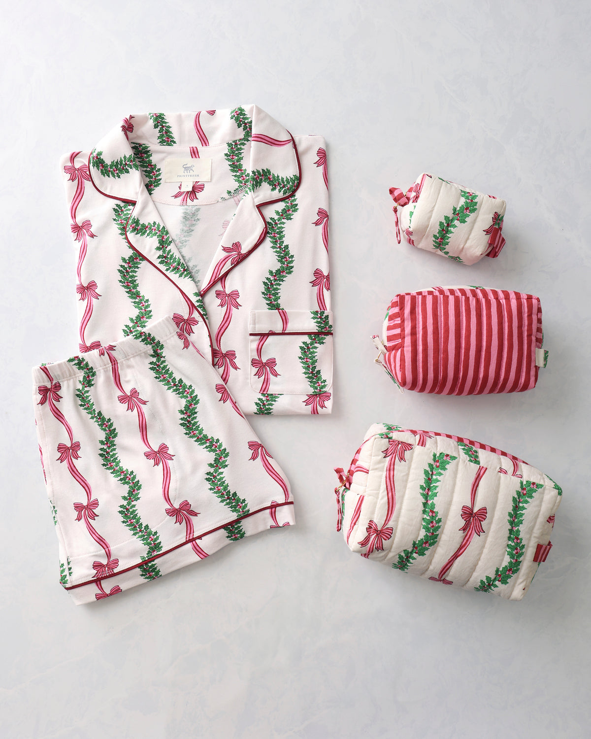 Folded white pajama top and shorts with red bow and green garland print beside three matching quilted pouches.