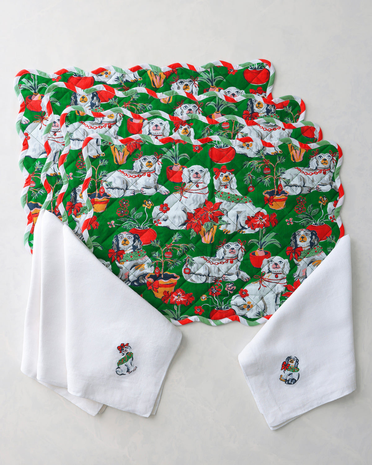 Four green placemats and white dinner napkins arranged together, featuring a Christmas pattern with dogs, bows, flowers, and holiday greenery.