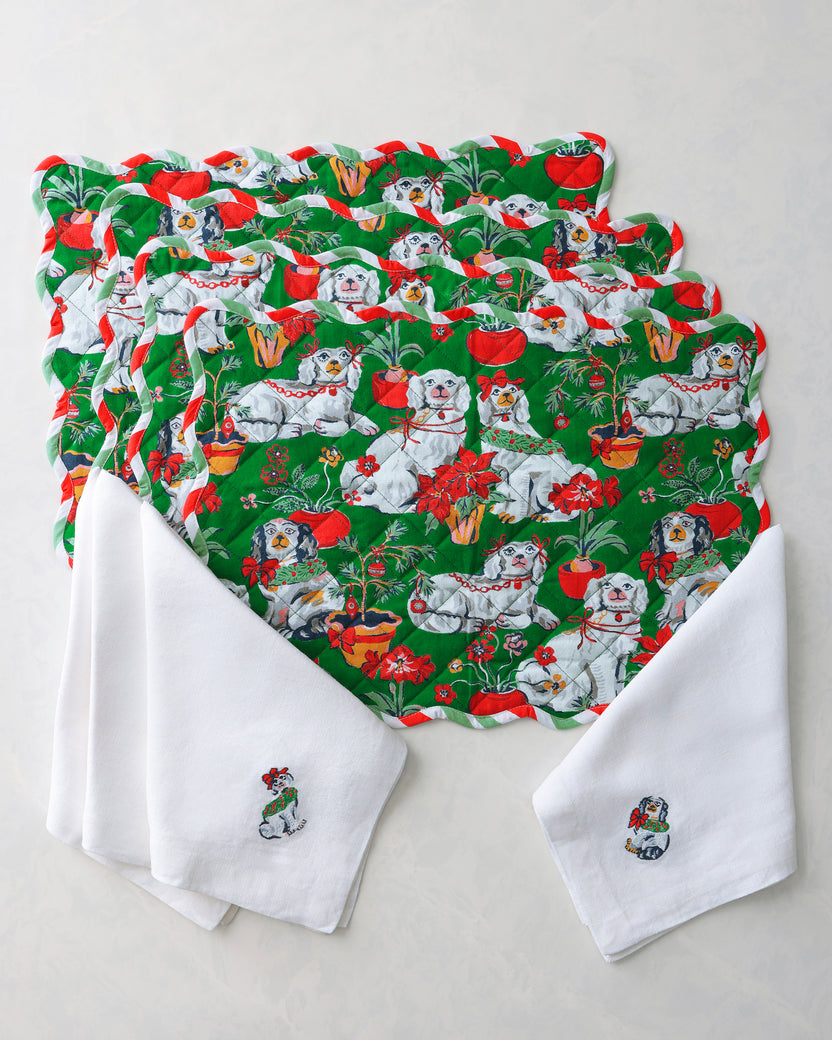 Four green placemats and white dinner napkins arranged together, featuring a Christmas pattern with dogs, bows, flowers, and holiday greenery.