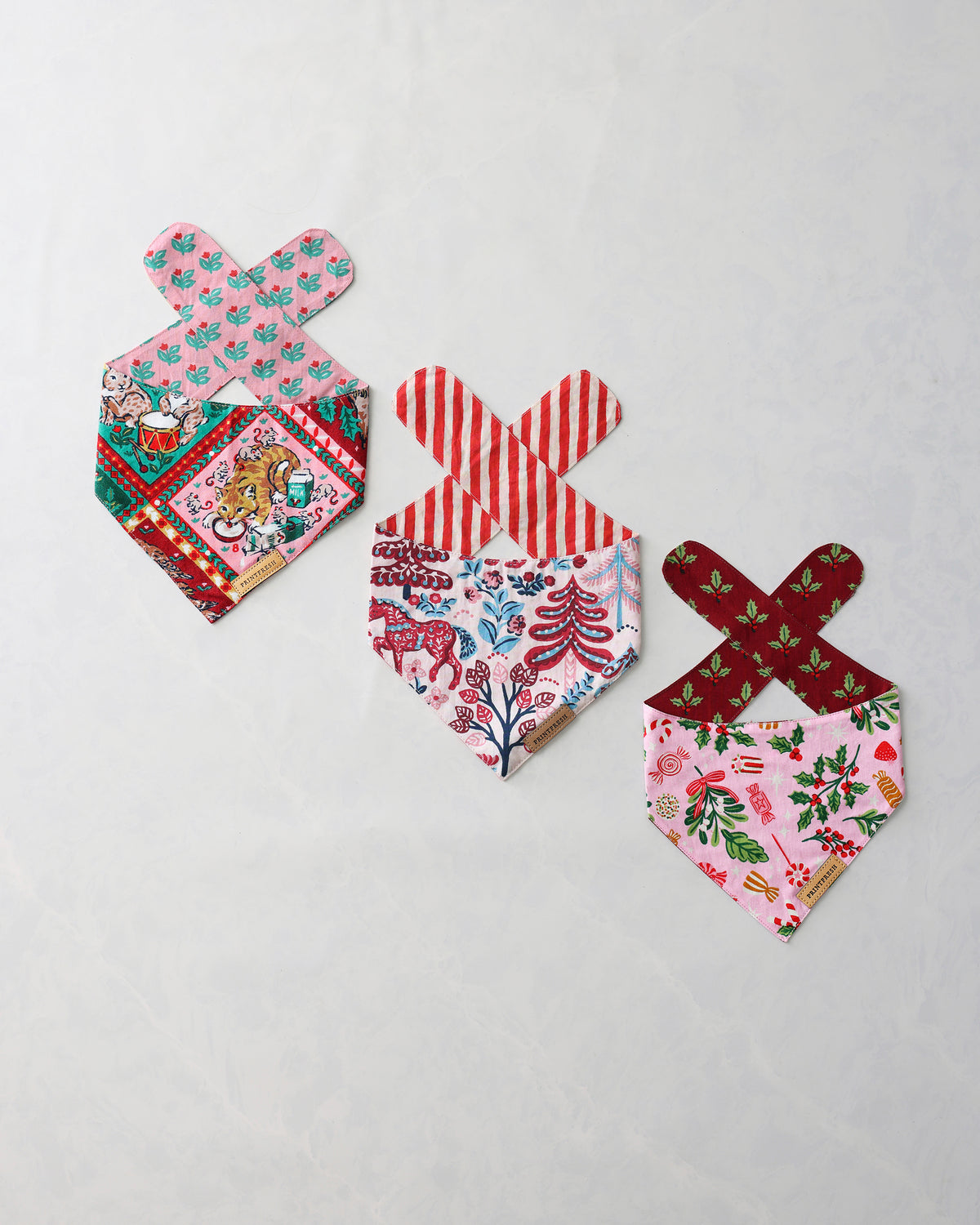 Three holiday pet bandanas arranged on a light background, with pink, red, green, and multicolored printed patterns and crossed tie straps.