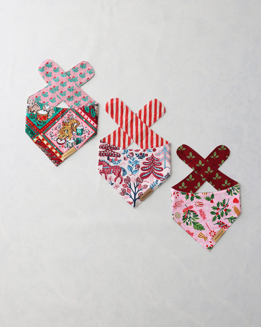 Three holiday pet bandanas arranged on a light background, with pink, red, green, and multicolored printed patterns and crossed tie straps.