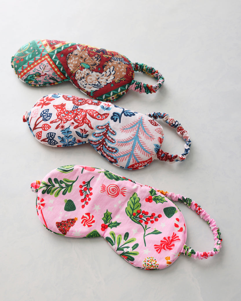 Three printed sleep masks arranged diagonally, featuring a red-and-green holiday pattern, a multicolored woodland pattern, and a pink candy-and-holly pattern.