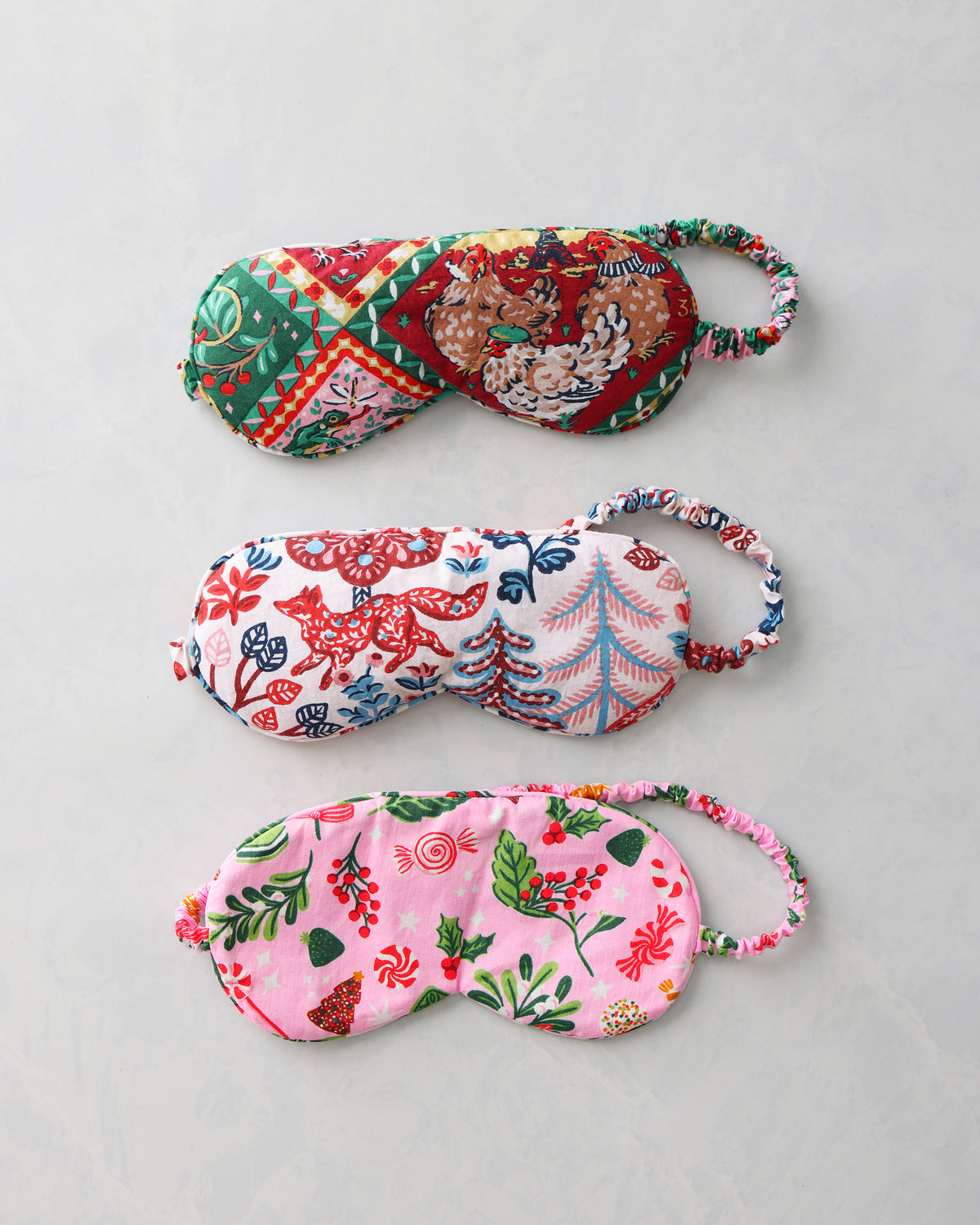 Three printed sleep masks shown separately on a light background, displaying their red-and-green, multicolored woodland, and pink holiday patterns.