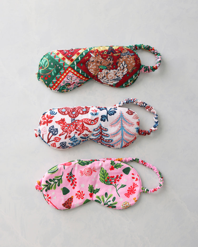 Three printed sleep masks shown separately on a light background, displaying their red-and-green, multicolored woodland, and pink holiday patterns.