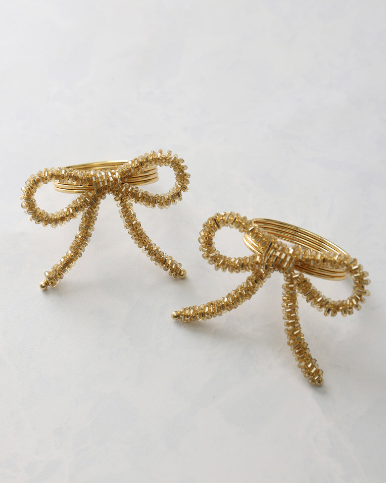 Two gold beaded bow napkin rings displayed on a light background.