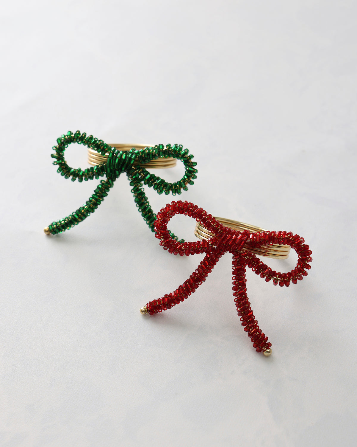 Two bow-shaped napkin rings with beaded detailing, one green and one red, each with a gold-tone metal center and dangling beaded tails.