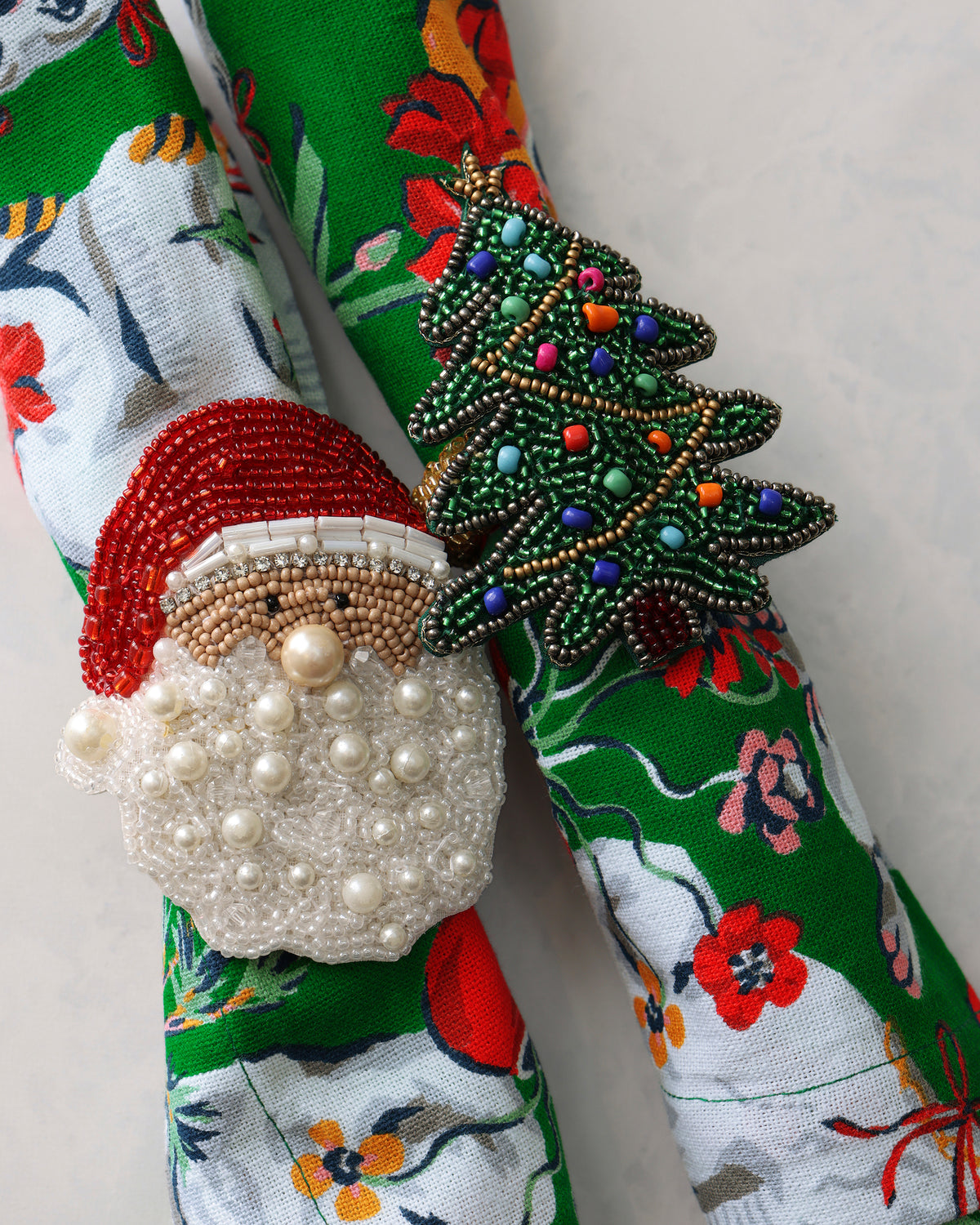 Two beaded holiday napkin rings, shaped as a Christmas tree and Santa, shown on patterned napkins.