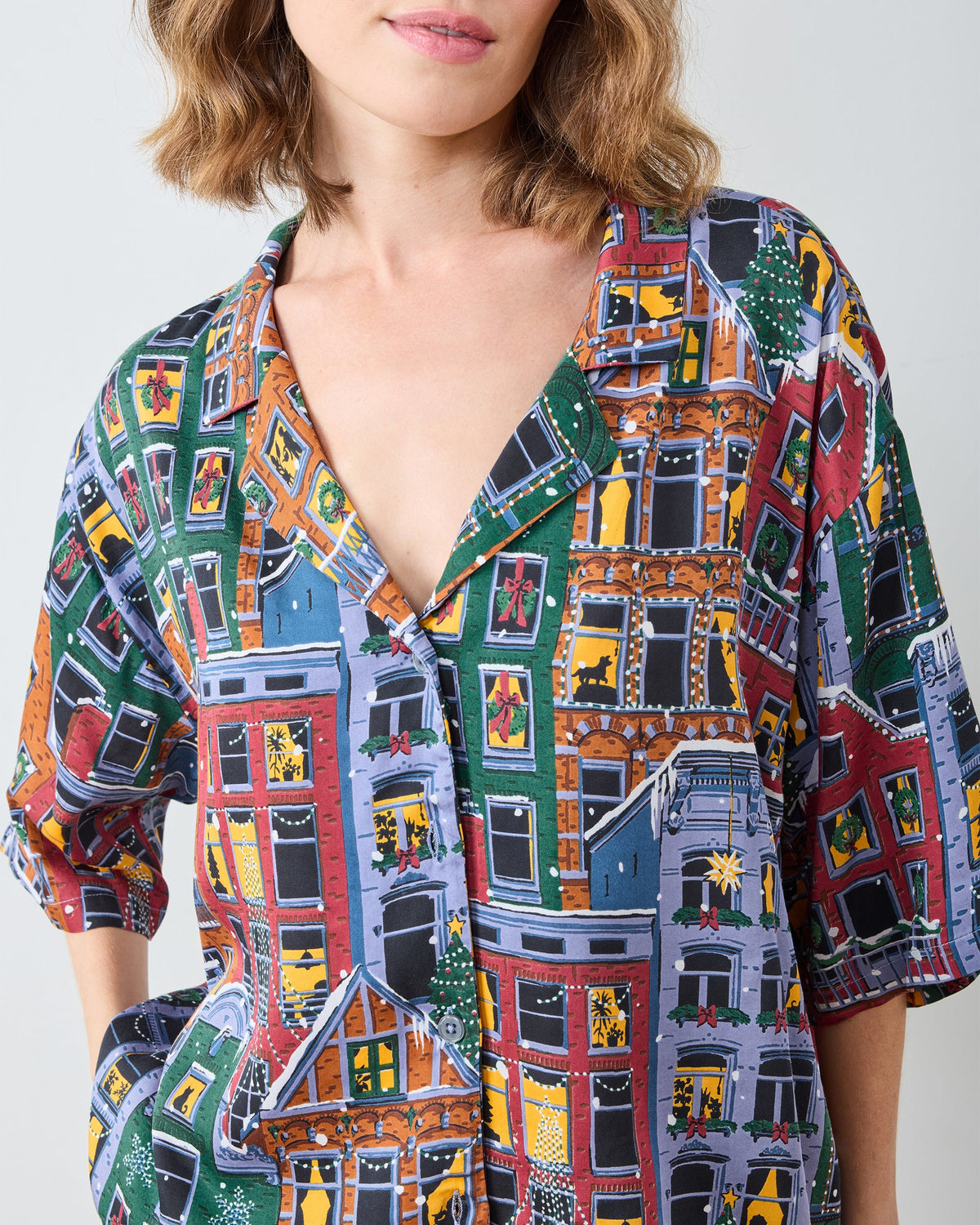 Close-up of the patterned pajama top showing its V-neck, button closure, short sleeves, illustrated buildings, windows, wreaths, and holiday trees.