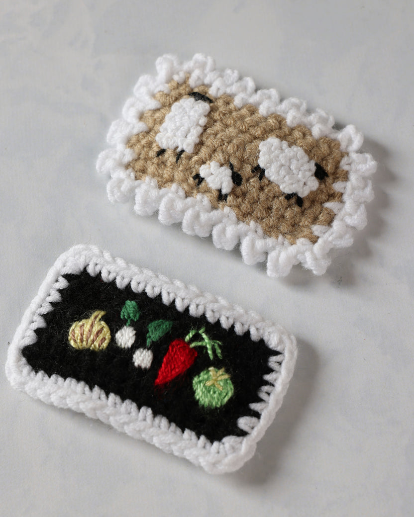 Angled close-up of two crochet snap hair clips with a sheep design and embroidered vegetables.
