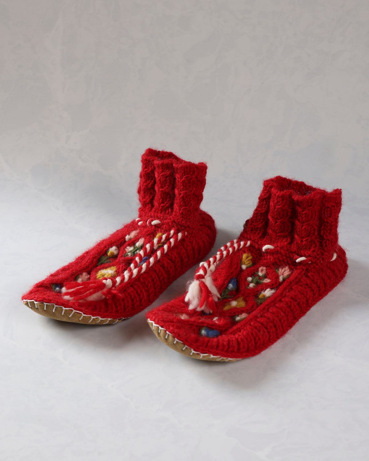 Pair of red handknit slippers with embroidered floral details, white-and-red ties, and leather soles, shown at an angle.