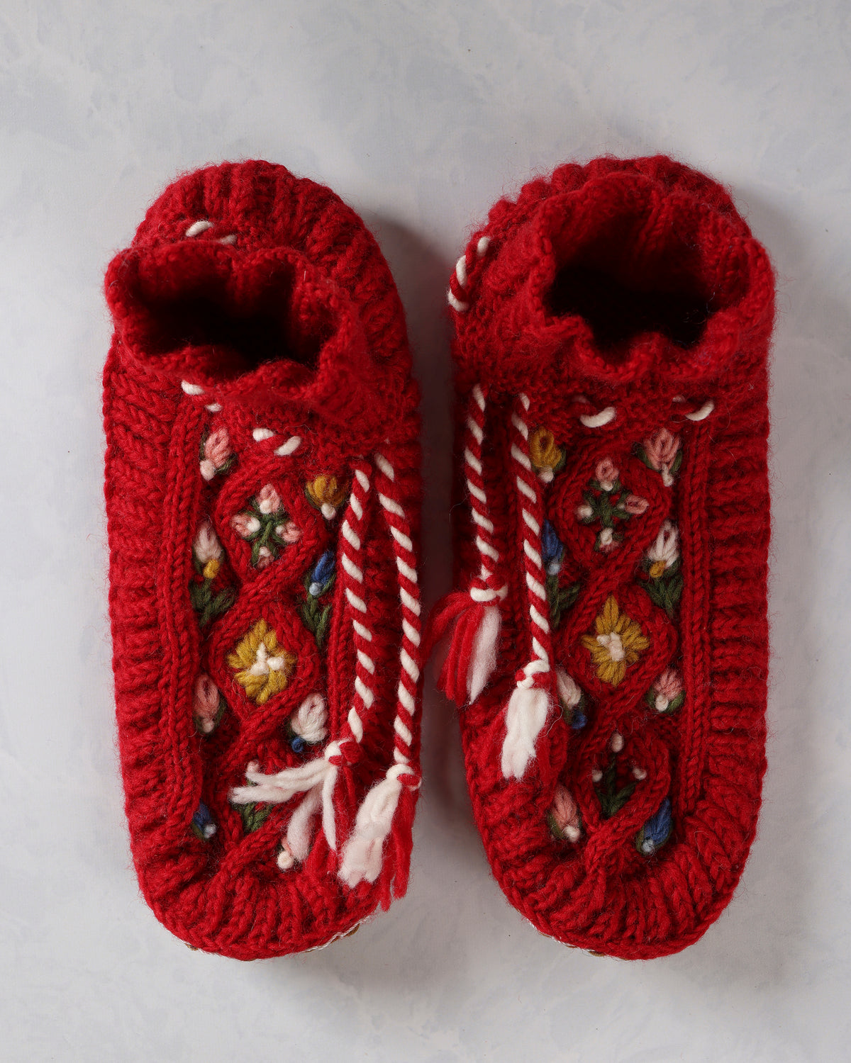 Top view of a pair of red handknit slippers with embroidered floral details and white-and-red ties.