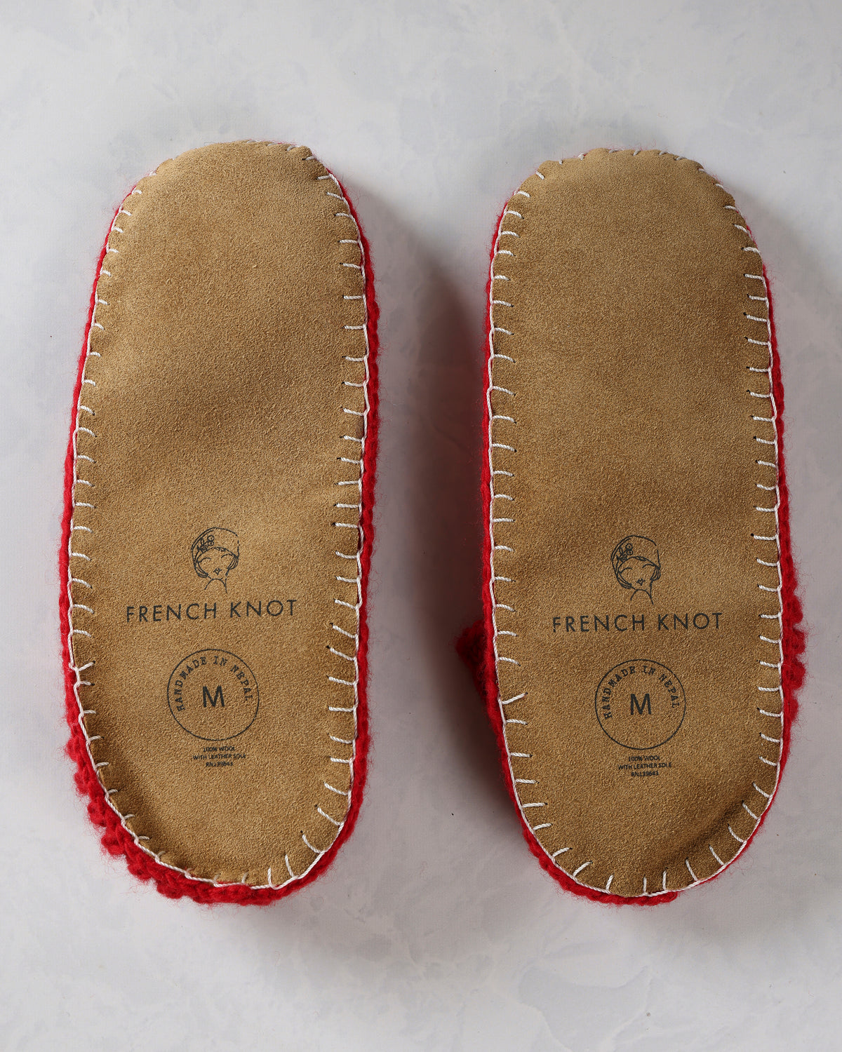 Bottom view of the pair of slippers showing tan leather soles with stitched edges and French Knot markings.