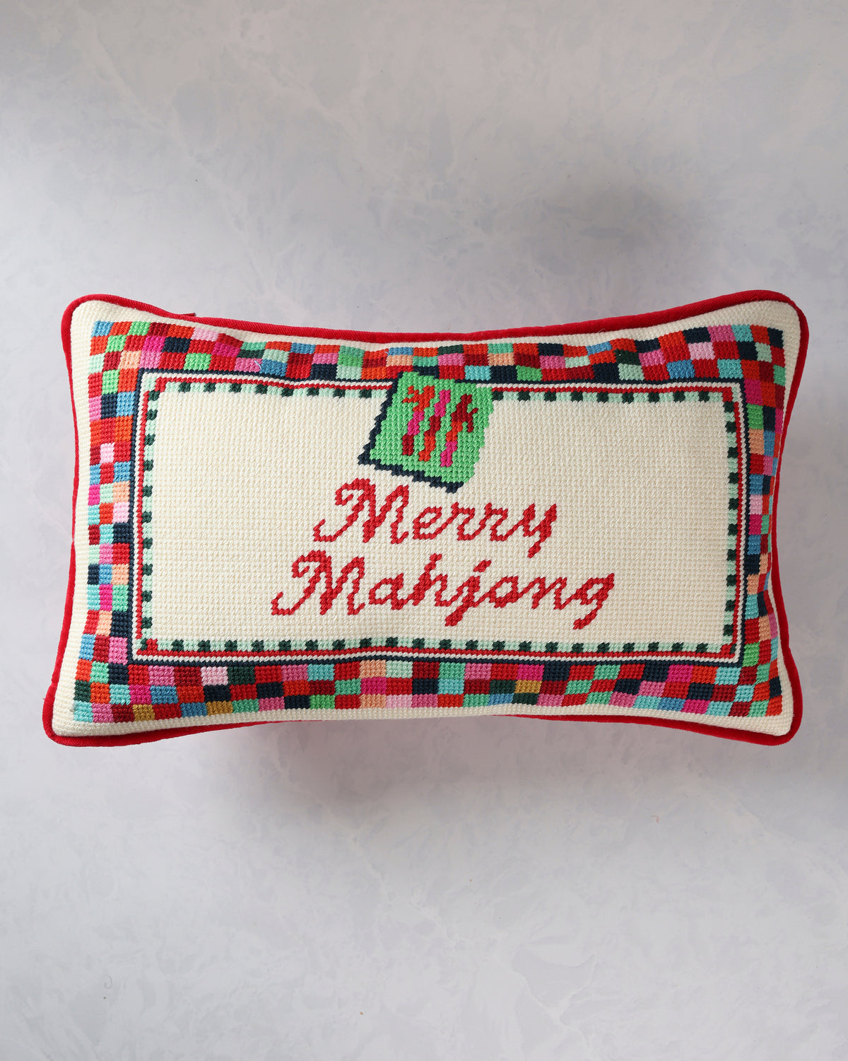 Rectangular needlepoint pillow with a multicolor border, green ornament motif, and red script reading “Merry Mahjong.”