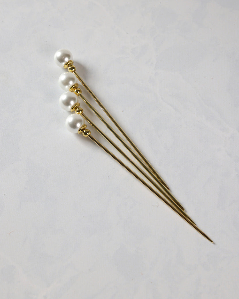 Four gold cocktail picks with faux pearl tops arranged diagonally.