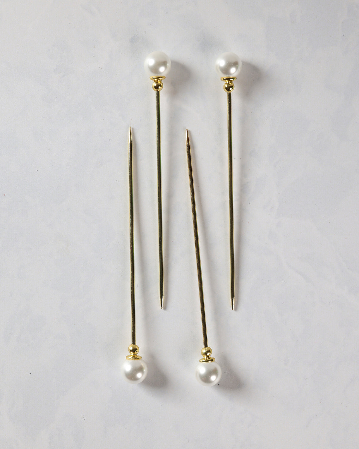 Four gold cocktail picks with pearl tops shown in two pairs, with pointed ends visible.