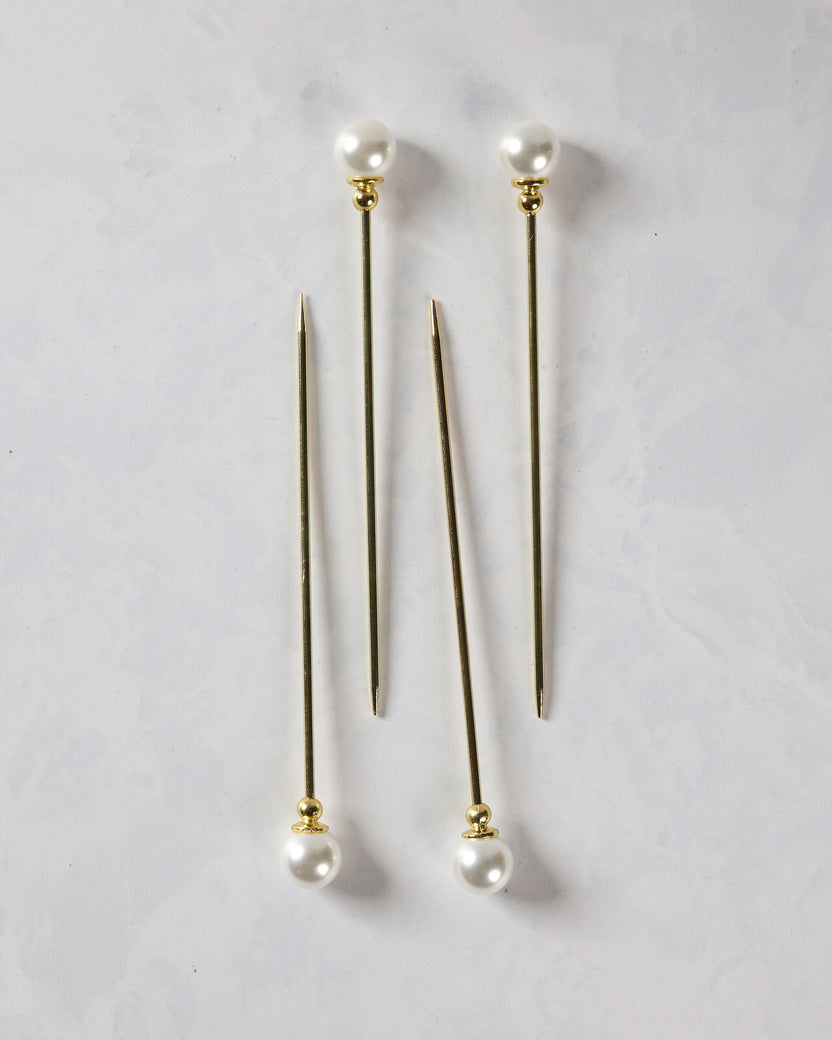 Four gold cocktail picks with pearl tops shown in two pairs, with pointed ends visible.
