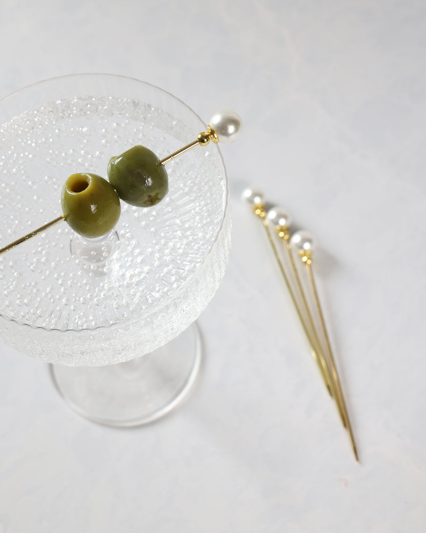 Gold cocktail pick with a pearl top shown beside green olives on a stemmed glass, with more pearl picks in the background.