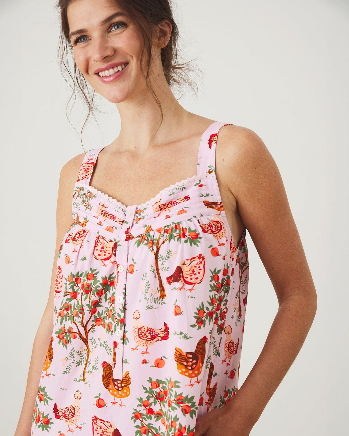 Front upper-body view of a ballet-pink nightgown showing its wide straps, scalloped neckline trim, white buttons, and hen-and-fruit print.