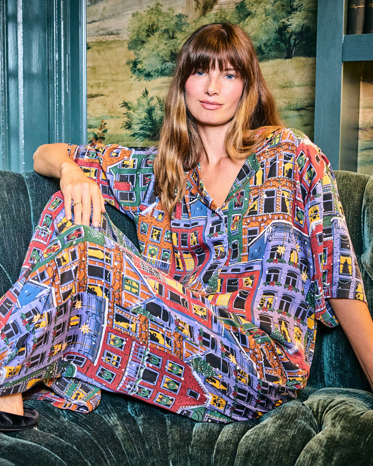 Person seated on a sofa wearing a colorful patterned pajama top and pants with illustrated city buildings and holiday details.