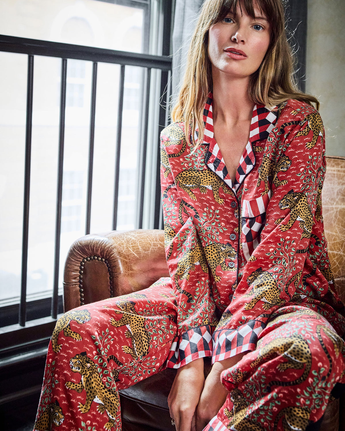 Model seated in a chair wearing a red leopard-print pajama shirt and pants with checkerboard trim.
