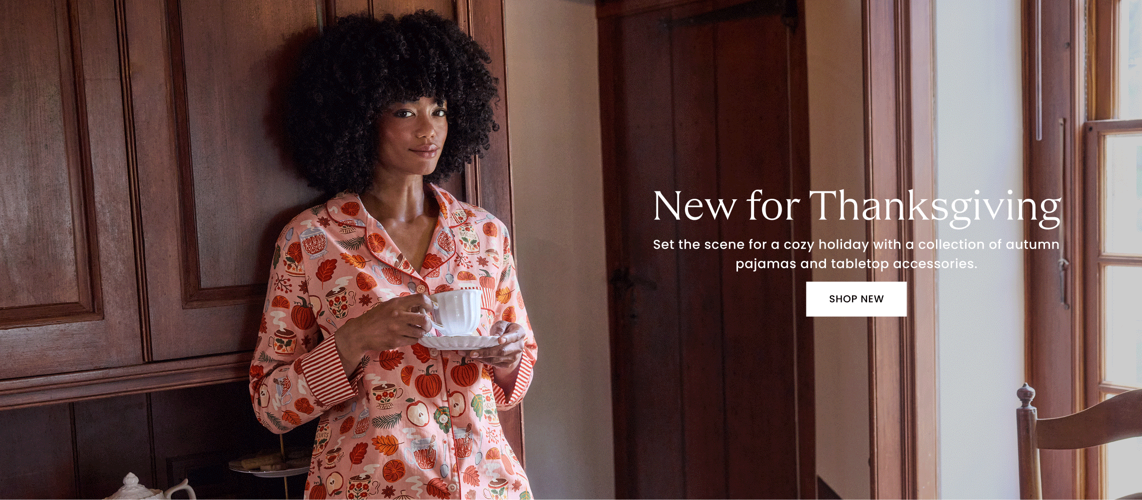 Wide promotional banner showing a person in pink holiday-print pajamas holding a cup, with text about new Thanksgiving pajamas and tabletop accessories and a SHOP NEW button.
