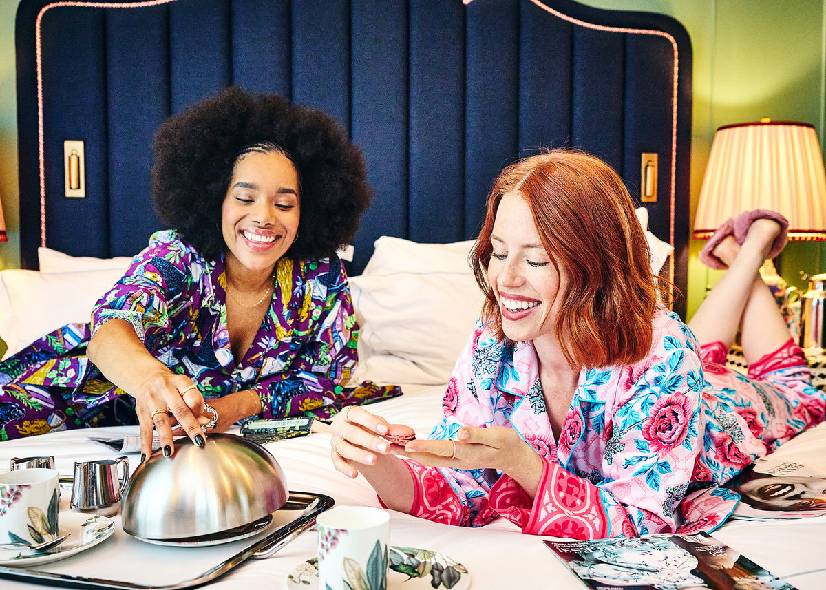 Two people in patterned pajamas sit on a bed beside a breakfast tray, cups, and a table lamp. 