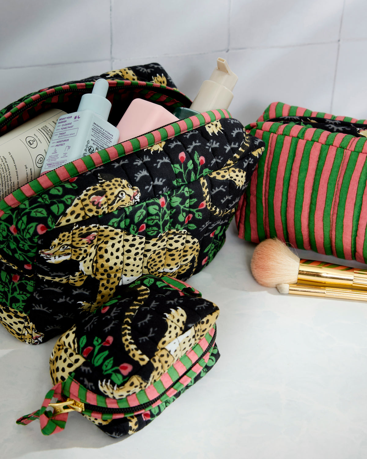 Three-piece Bagheera pouch set arranged with an open leopard-print pouch, a striped pouch, and a smaller zip pouch beside makeup items and a brush.