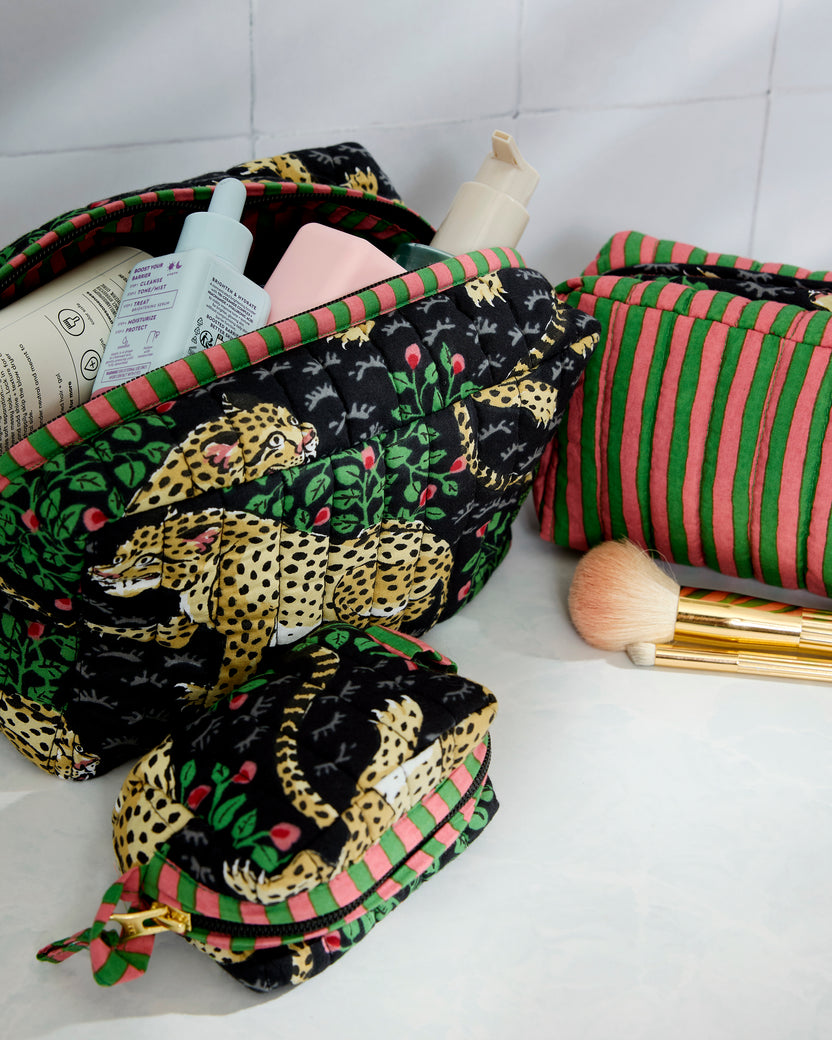 Three-piece Bagheera pouch set arranged with an open leopard-print pouch, a striped pouch, and a smaller zip pouch beside makeup items and a brush.