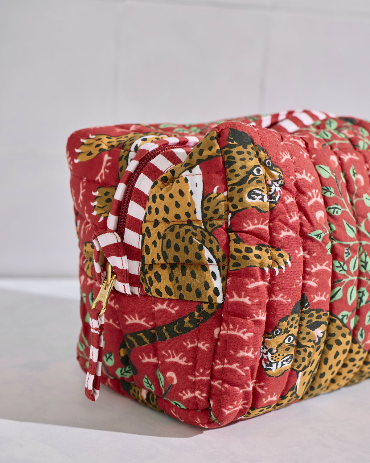 Angled close view of a red quilted Bagheera pouch showing leopard motifs, green leaves, and a red-and-white striped zipper trim.