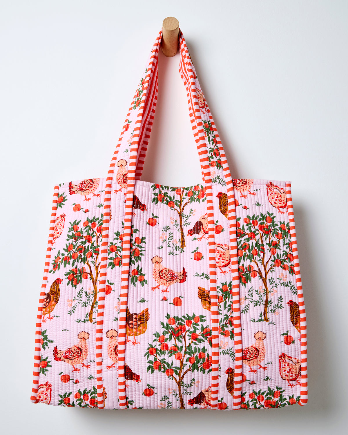 Quilted tote bag hanging against a wall, with a light pink farmyard print, striped handles, chickens, and fruit trees.