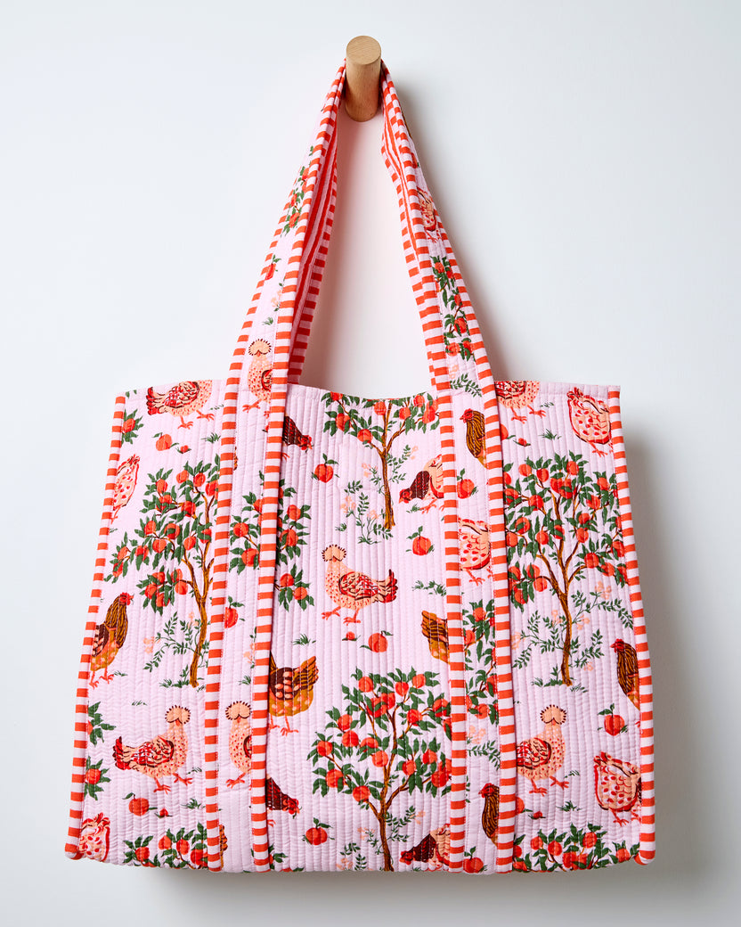 Quilted tote bag hanging against a wall, with a light pink farmyard print, striped handles, chickens, and fruit trees.