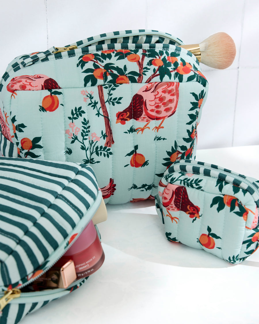 Three quilted pouches in coordinated soft blue prints, including farm animals, fruit, and stripes; one pouch is open with makeup items visible.