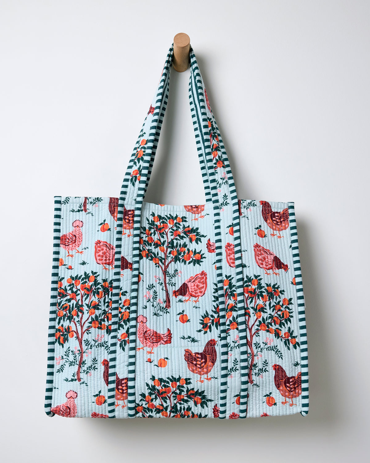 Quilted tote bag hanging against a wall, with a light blue farmyard print, striped handles, chickens, and fruit trees.