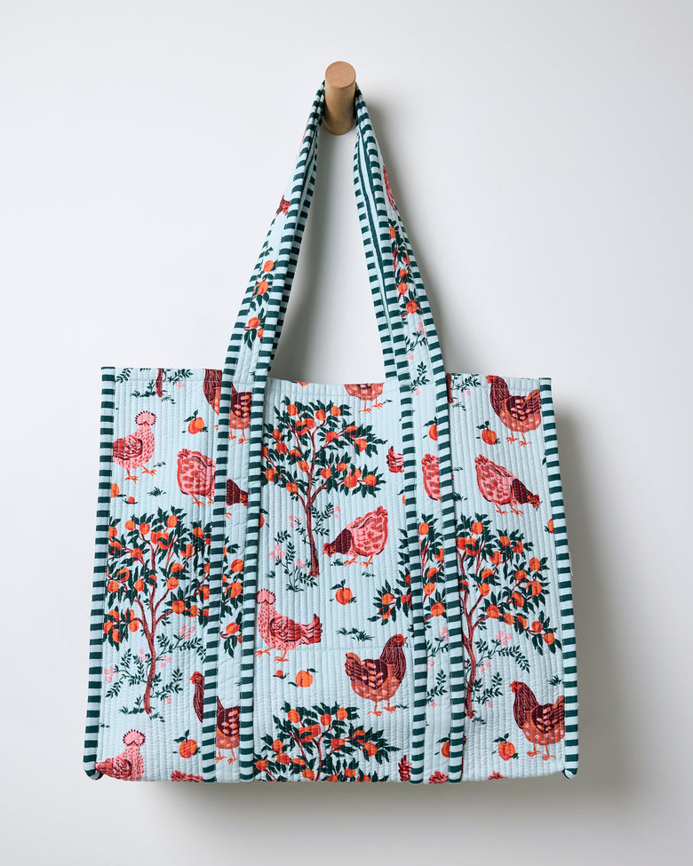Quilted tote bag hanging against a wall, with a light blue farmyard print, striped handles, chickens, and fruit trees.