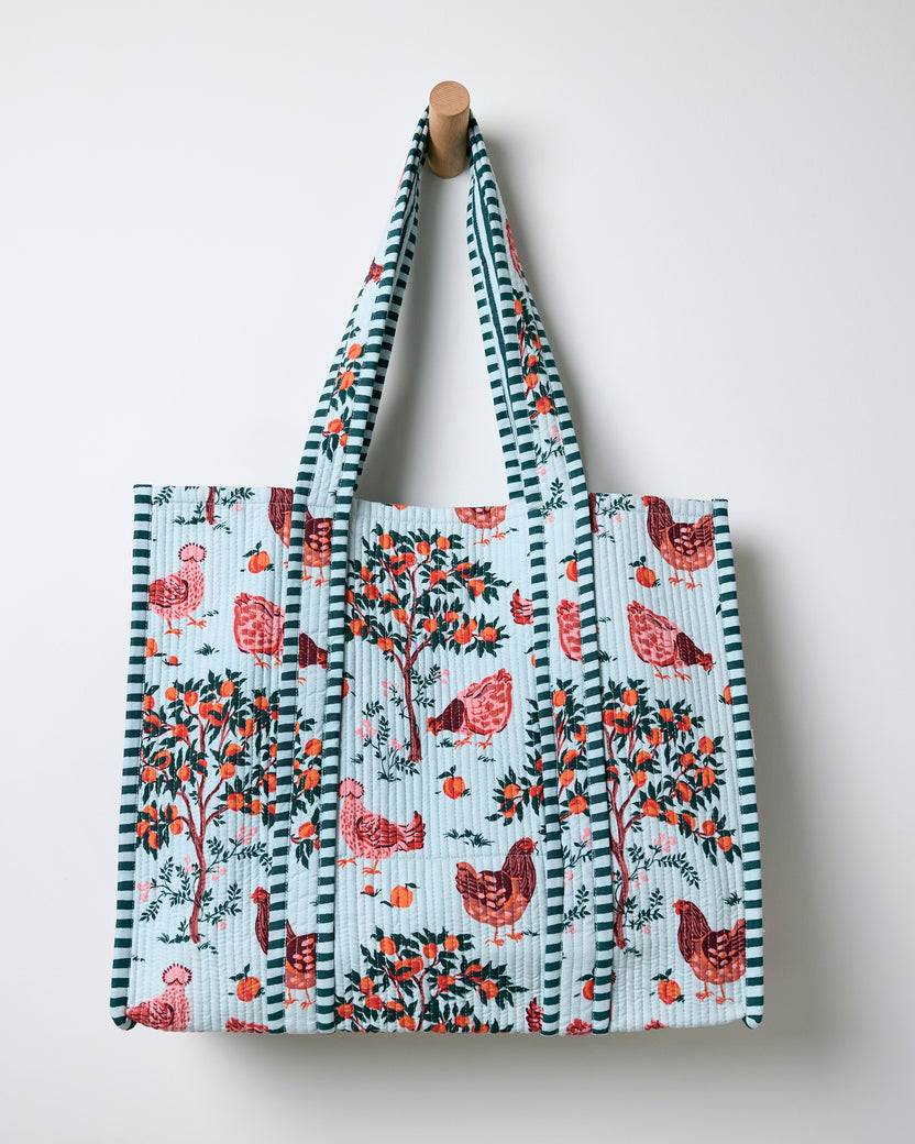 Quilted tote bag hanging against a wall, with a light blue farmyard print, striped handles, chickens, and fruit trees.