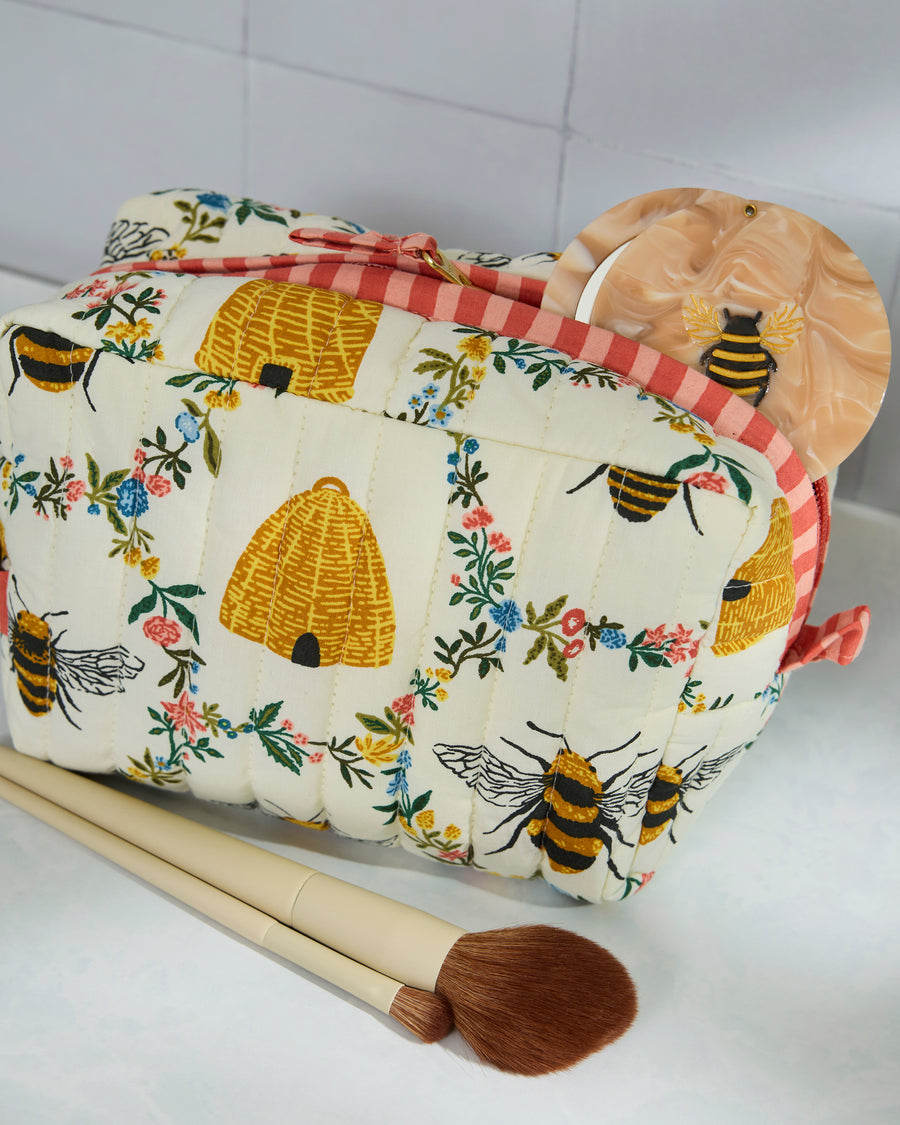 Cream quilted pouch with bees, beehives, and floral print, shown with makeup brushes and a round compact.