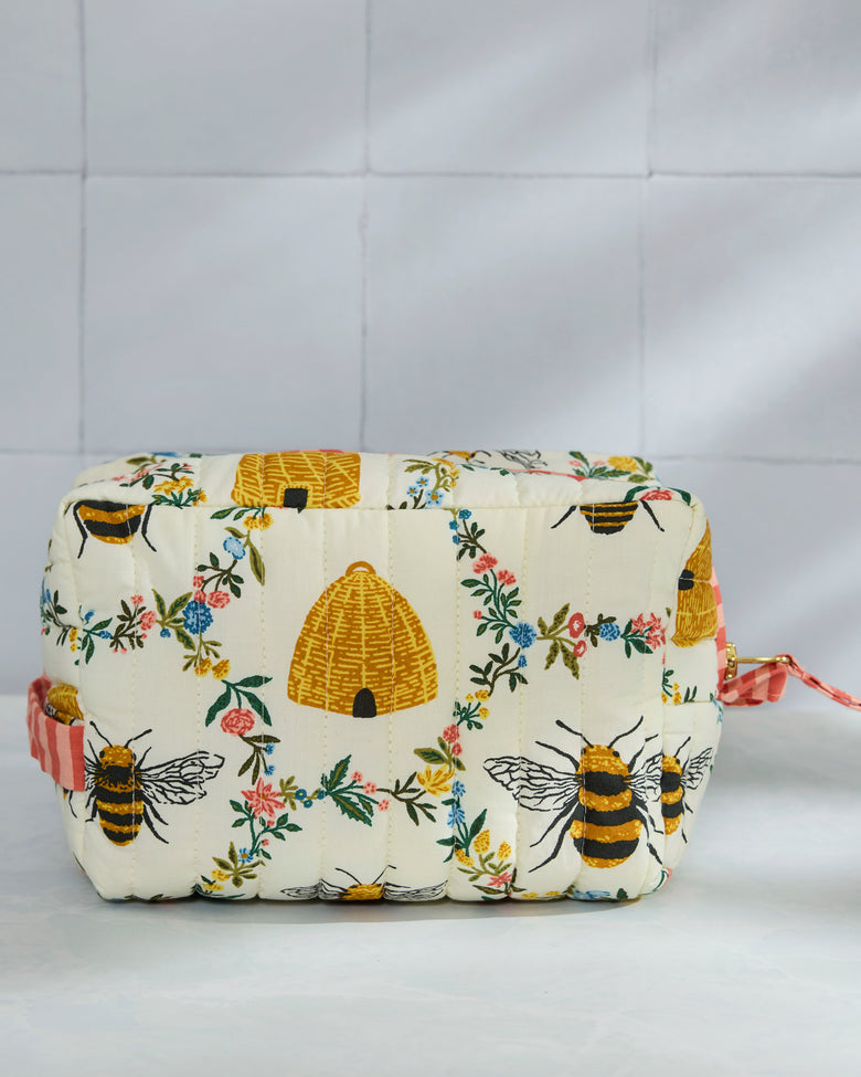 Front view of a cream quilted pouch with repeated bees, beehives, and floral print, plus pink striped trim and a side strap.