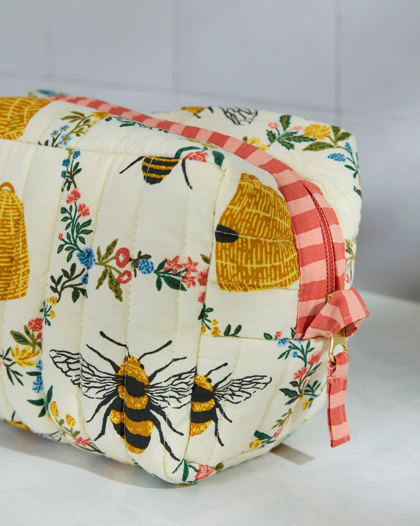 Close detail of the cream quilted pouch showing bees, beehives, floral print, pink striped zipper trim, and side strap hardware.