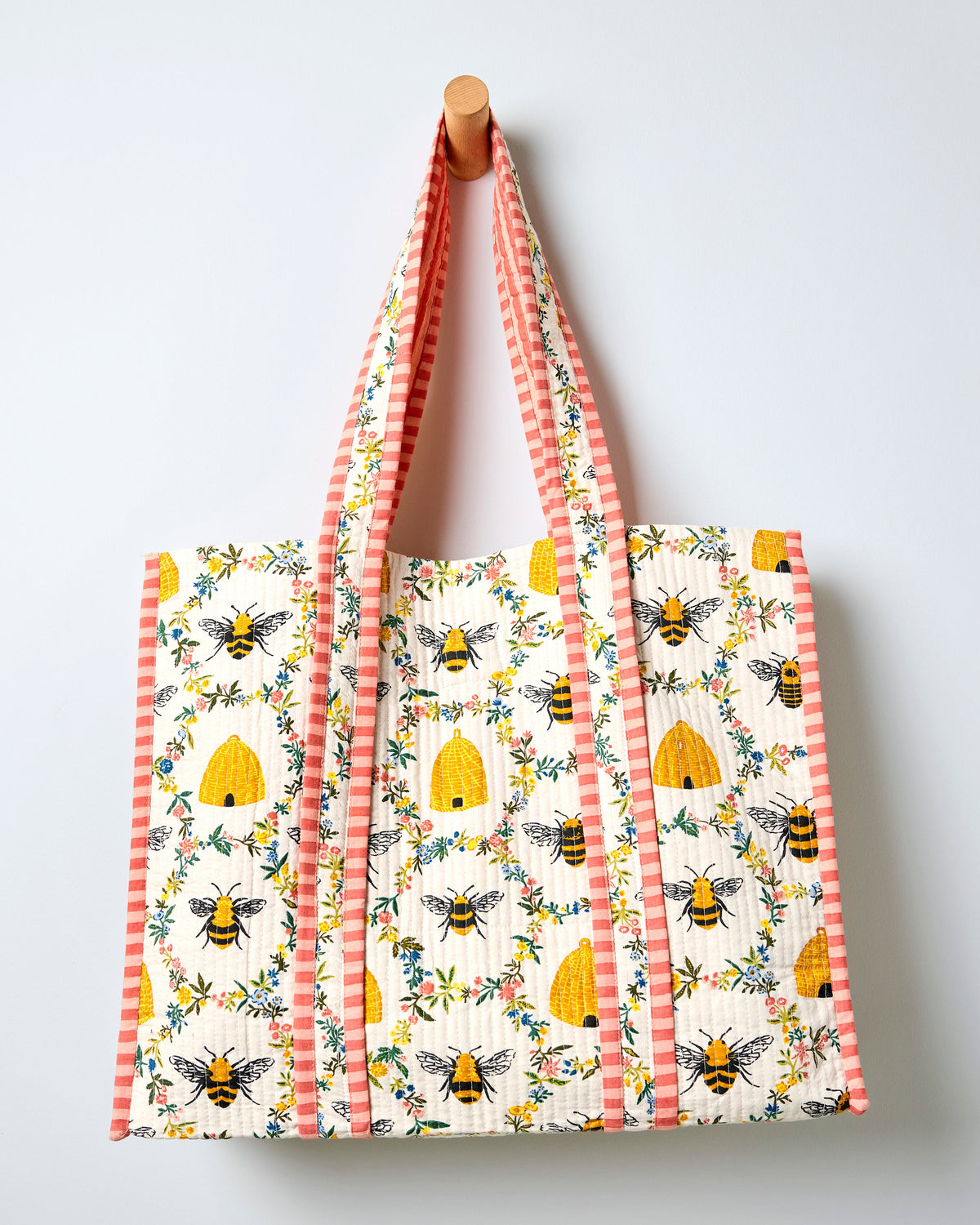 Full view of a cream quilted traveler tote with repeated bee, beehive, and floral print, framed by pink striped handles and trim.